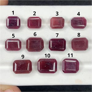 May include: Eleven rectangular, faceted, deep red rubies arranged in three rows on a white display tray. Each gemstone is individually numbered from one to eleven in black text, showcasing varying shades of crimson and distinct internal inclusions.