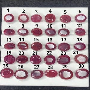 May include: A collection of thirty oval-cut, deep pink to red gemstones arranged in five rows of six on a white display tray. Each gemstone is individually numbered from one to thirty, featuring faceted surfaces that reflect light.
