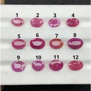 Pode incluir: Doze pedras preciosas ovais e facetadas, de cor rosa e vermelha, dispostas em três filas numa bandeja de apresentação branca. Cada pedra está marcada com um número preto de 1 a 12, exibindo vários tons de rosa e vermelho translúcido.