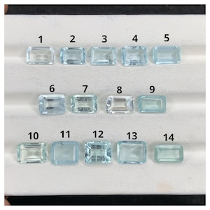 Puede incluir: Catorce piedras preciosas de aguamarina rectangulares de varios tonos de azul claro y transparente. Las piedras están dispuestas en filas sobre una superficie blanca, cada una con un corte y claridad únicos. Numeradas del 1 al 14.