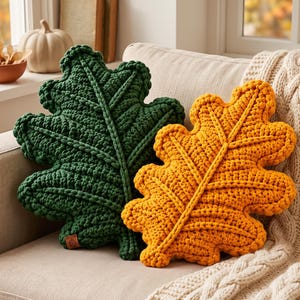 Peut inclure: Deux coussins décoratifs en forme de feuilles de chêne, l'un vert forêt profond et l'autre jaune doré vif. Les deux coussins présentent un motif en maille crochetée épaisse et texturée avec un détail de nervure centrale bien visible.
