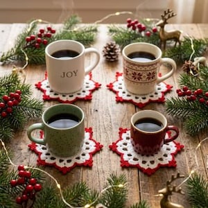 May include: Four ceramic mugs filled with dark coffee, each resting on a white and red crocheted snowflake coaster. One mug is white with the text JOY, one has a red and white Nordic pattern, one is light green, and one is solid dark red.