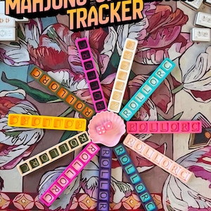 May include: A Mahjong Charleston Tracker set featuring ten colorful rectangular plastic bars with the text ROLLORC, arranged in a circle around a small pink shell dish containing two pink dice. The text MAHJONG CHARLESTON TRACKER and PICK YOUR COLORS is visible.