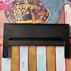 Puede incluir: Un soporte de pl&aacute;stico negro con un marco rectangular y una barra de soporte horizontal. El soporte presenta una superficie s&oacute;lida y lisa con dos patas verticales que se extienden hacia abajo, dise&ntilde;ado para sostener objetos en posici&oacute;n vertical.