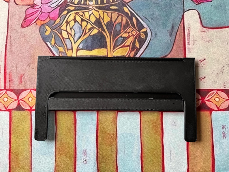 Puede incluir: Un soporte de pl&aacute;stico negro con un marco rectangular y una barra de soporte horizontal. El soporte presenta una superficie s&oacute;lida y lisa con dos patas verticales que se extienden hacia abajo, dise&ntilde;ado para sostener objetos en posici&oacute;n vertical.