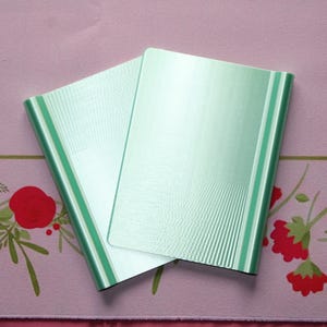 May include: Two rectangular, light mint green folders with a subtle, fine-lined vertical texture. The folders are positioned overlapping each other, displaying a smooth, metallic-like finish on their front covers and rounded edges along the sides.