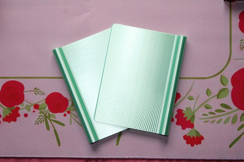May include: Two rectangular, light mint green folders with a subtle, fine-lined vertical texture. The folders are positioned overlapping each other, displaying a smooth, metallic-like finish on their front covers and rounded edges along the sides.