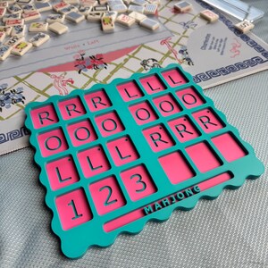 May include: A teal and pink Mahjong card holder featuring a grid of recessed squares with the letters R, L, and O, and the numbers 1, 2, and 3 embossed in the teal frame. The word MAHJONG is printed in teal on a horizontal bar at the bottom.