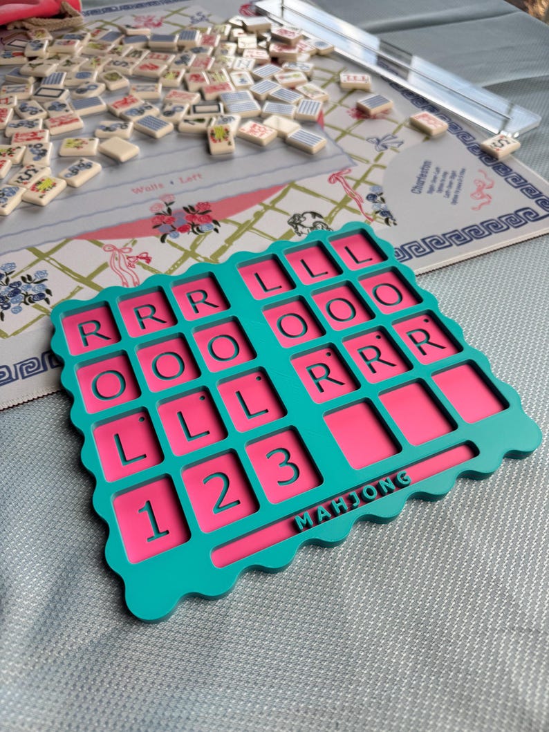 May include: A teal and pink Mahjong card holder featuring a grid of recessed squares with the letters R, L, and O, and the numbers 1, 2, and 3 embossed in the teal frame. The word MAHJONG is printed in teal on a horizontal bar at the bottom.