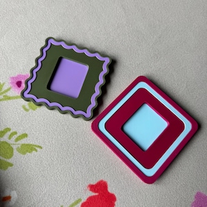 May include: Two colorful, geometric coasters. One is olive green with a purple center and a scalloped edge. The other is a maroon and light blue diamond shape. The coasters are on a floral patterned surface.