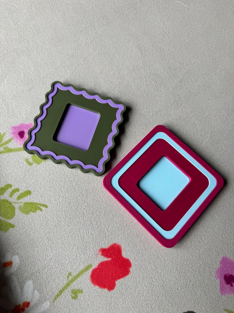 May include: Two colorful, geometric coasters. One is olive green with a purple center and a scalloped edge. The other is a maroon and light blue diamond shape. The coasters are on a floral patterned surface.