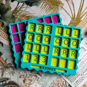 May include: Two teal plastic Mahjong organizers with scalloped edges. The top tray features a lime green interior with grid compartments labeled with the letters R, O, L, and the numbers 1, 2, 3. The word MAHJONG is embossed at the bottom center.