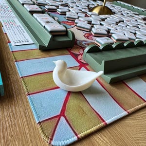 May include: A white bird-shaped dish sits on a patterned mat featuring light blue, olive green, and red geometric lines. Behind it are green tile racks and a collection of white mahjong tiles with various symbols, alongside a scoring card.