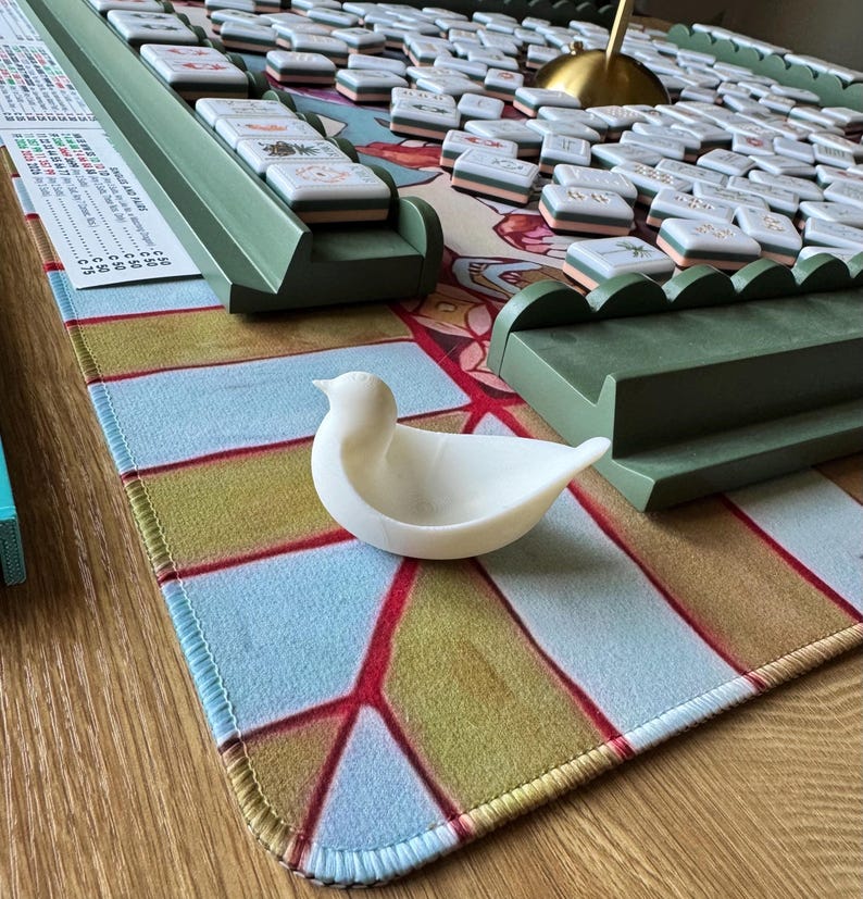 May include: A white bird-shaped dish sits on a patterned mat featuring light blue, olive green, and red geometric lines. Behind it are green tile racks and a collection of white mahjong tiles with various symbols, alongside a scoring card.