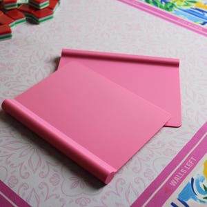May include: Two solid pink rectangular mahjong tile racks with a smooth matte finish and a rounded top edge. The racks are positioned at an angle, overlapping each other, and feature a simple, clean design for holding game tiles during play.