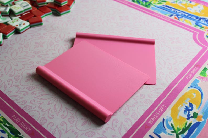 May include: Two solid pink rectangular mahjong tile racks with a smooth matte finish and a rounded top edge. The racks are positioned at an angle, overlapping each other, and feature a simple, clean design for holding game tiles during play.