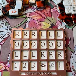 May include: A rose gold colored plastic tray with a scalloped edge, featuring a grid of twenty-four square slots containing tiles with letters R, O, L, numbers 1, 2, 3, a lotus flower, a bird, and a bamboo stalk. The text MAHJONG BAM is embossed.