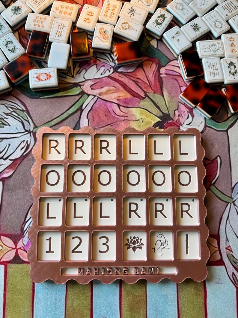 May include: A rose gold colored plastic tray with a scalloped edge, featuring a grid of twenty-four square slots containing tiles with letters R, O, L, numbers 1, 2, 3, a lotus flower, a bird, and a bamboo stalk. The text MAHJONG BAM is embossed.