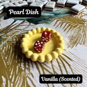 May include: A round yellow dish with a textured beaded rim containing two translucent red dice with white pips. The dish is labeled Pearl Dish and Vanilla (Scented). In the background are numerous white rectangular tiles with various black and colored symbols.