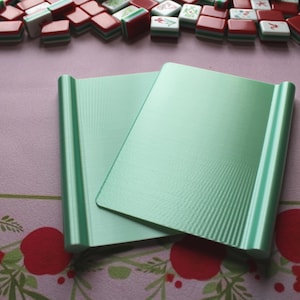 May include: Two rectangular, light mint green plastic mahjong tile racks with a smooth, flat central surface and rounded, raised edges on both sides. The racks are positioned overlapping each other, featuring a subtle vertical ribbed texture.