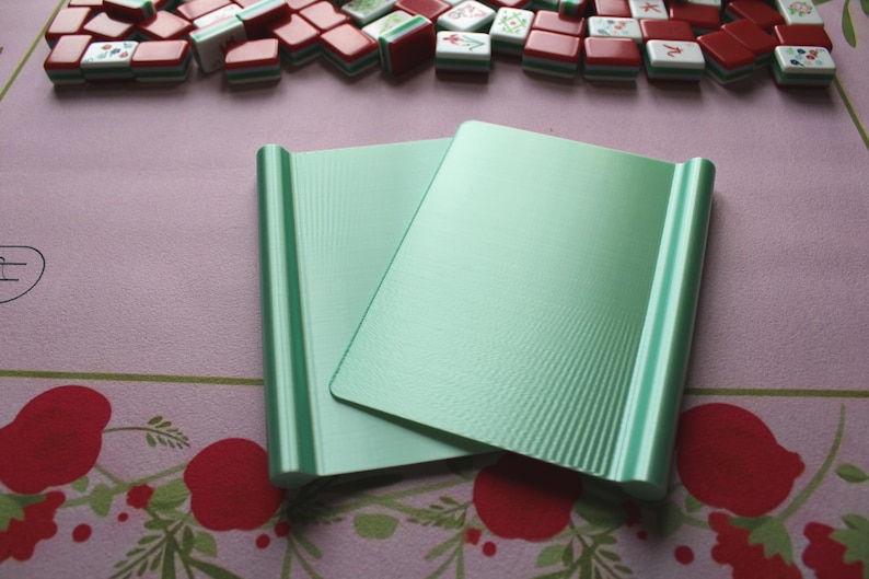 May include: Two rectangular, light mint green plastic mahjong tile racks with a smooth, flat central surface and rounded, raised edges on both sides. The racks are positioned overlapping each other, featuring a subtle vertical ribbed texture.