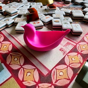 May include: A bright pink, bird-shaped bowl with a smooth, matte finish. The bowl features a curved, wing-like structure and a pointed tail, resting on a surface covered with scattered white mahjong tiles and a gold-colored lamp base.