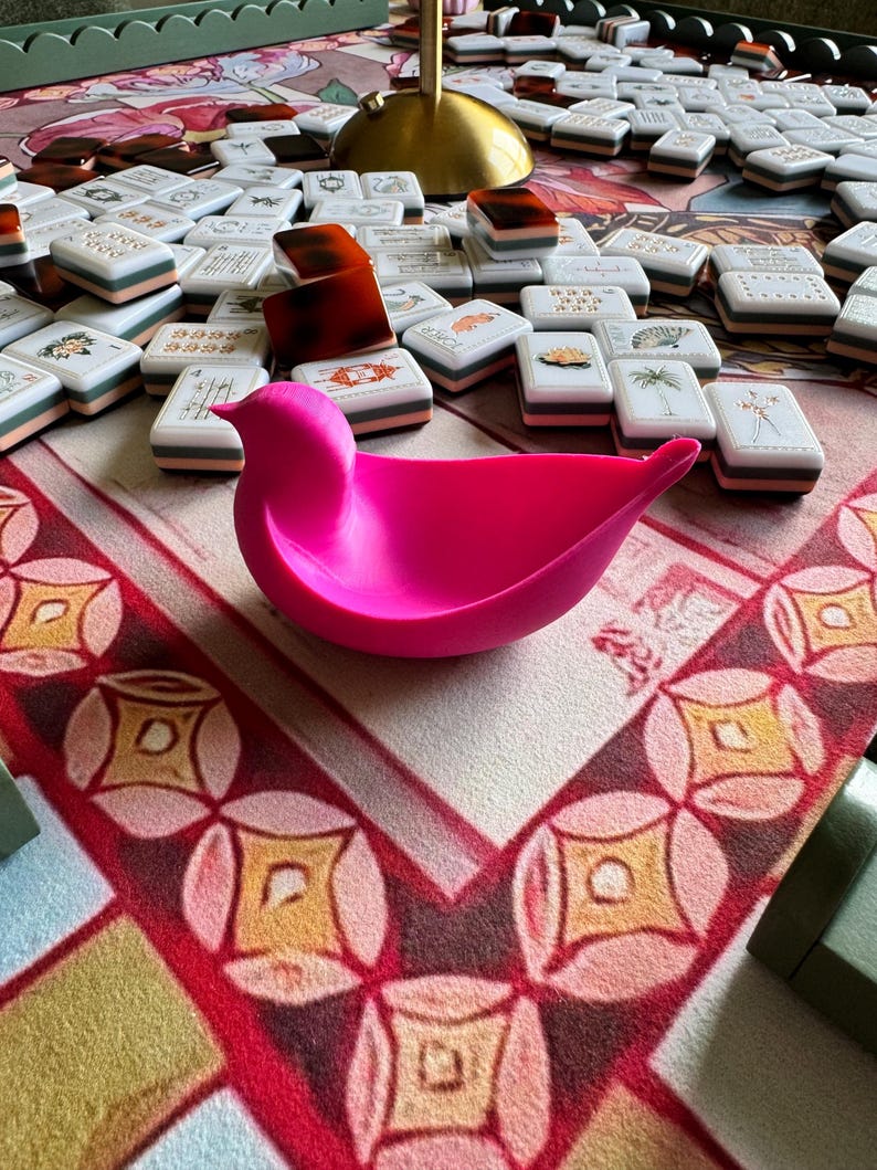 May include: A bright pink, bird-shaped bowl with a smooth, matte finish. The bowl features a curved, wing-like structure and a pointed tail, resting on a surface covered with scattered white mahjong tiles and a gold-colored lamp base.