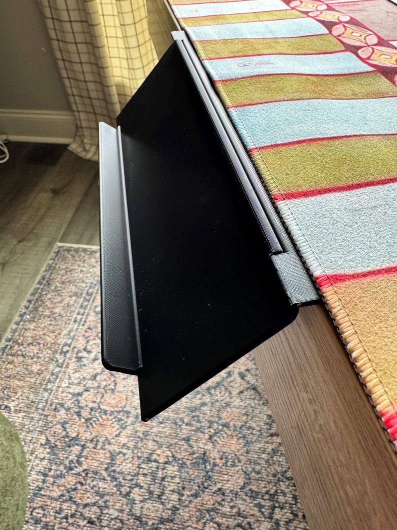 Puede incluir: Una bandeja de extensi&oacute;n de escritorio de pl&aacute;stico negro con una superficie plana y un borde frontal elevado, dise&ntilde;ada para fijarse al borde de una mesa. La bandeja presenta un acabado texturizado y un soporte de montaje resistente.