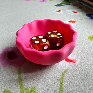 May include: A small, bright pink bowl with a scalloped, wavy rim. Inside the bowl are two translucent, amber-colored dice with white pips. The bowl has a matte finish and a rounded, shallow shape, contrasting with the glossy surface of the dice.