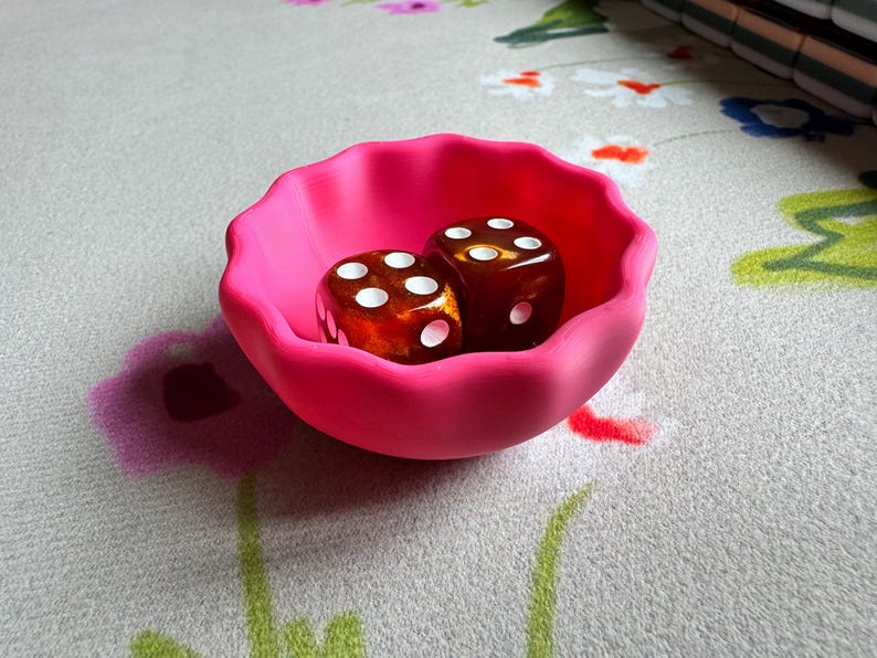 May include: A small, bright pink bowl with a scalloped, wavy rim. Inside the bowl are two translucent, amber-colored dice with white pips. The bowl has a matte finish and a rounded, shallow shape, contrasting with the glossy surface of the dice.