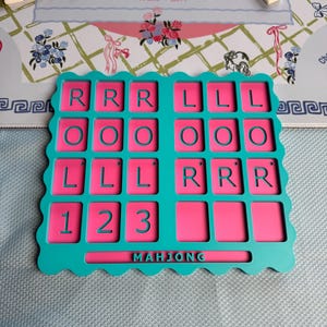 May include: A teal and pink plastic Mahjong tile rack with a scalloped edge. The rack features a grid of rectangular slots with embossed letters R, L, O, and numbers 1, 2, 3. The word MAHJONG is embossed in the center bottom section of the rack.