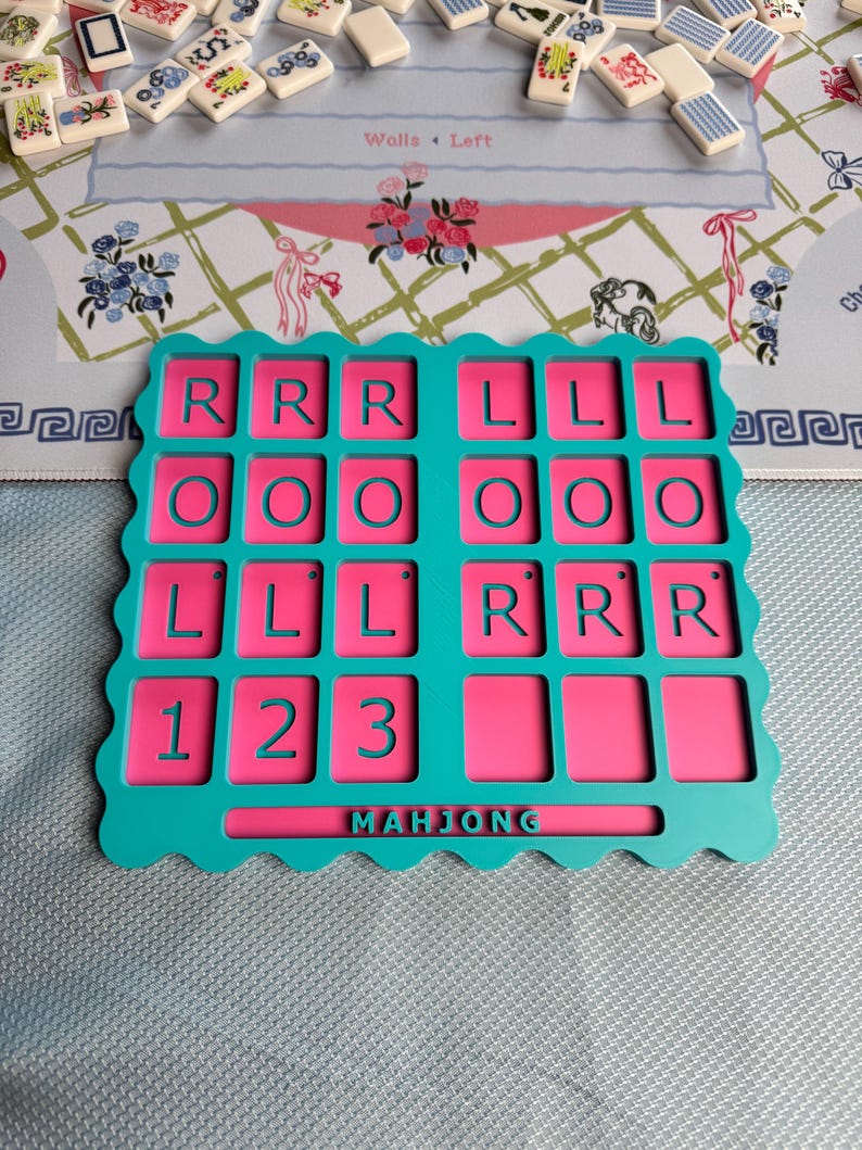 May include: A teal and pink plastic Mahjong tile rack with a scalloped edge. The rack features a grid of rectangular slots with embossed letters R, L, O, and numbers 1, 2, 3. The word MAHJONG is embossed in the center bottom section of the rack.