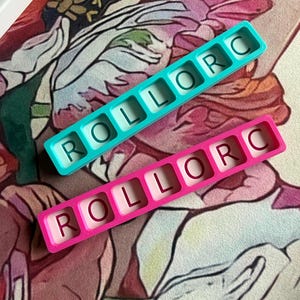 May include: Two rectangular silicone molds, one teal and one pink, each featuring six square cavities. Each cavity contains a single embossed letter, spelling out the word ROLLORC in a horizontal row within each mold.