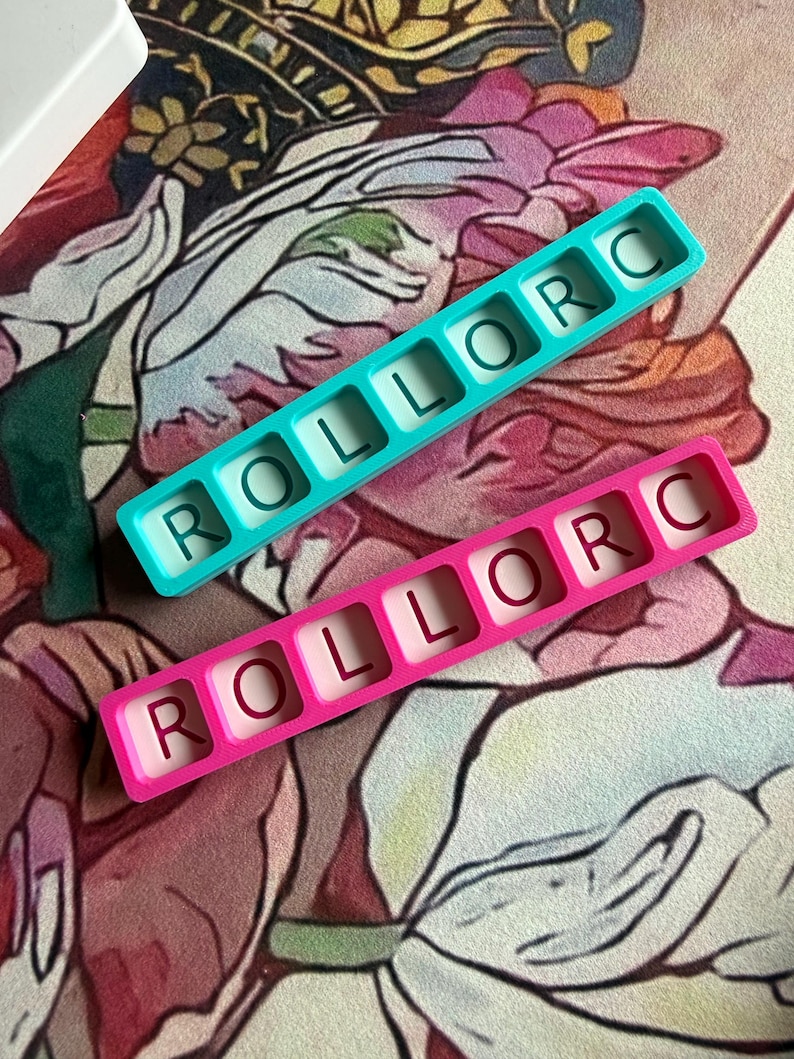 May include: Two rectangular silicone molds, one teal and one pink, each featuring six square cavities. Each cavity contains a single embossed letter, spelling out the word ROLLORC in a horizontal row within each mold.