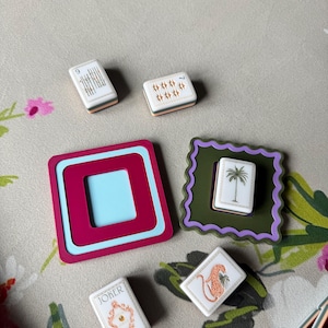 May include: A collection of decorative Mahjong tiles and coasters. The tiles feature various designs, including a palm tree, a leopard, and the word "Joker." The coasters are in shades of pink, blue, and green.