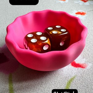 May include: A hot pink bowl with a scalloped edge containing two amber-colored dice with white pips. The image includes the text Bowl and Hot Pink. The bowl has a smooth, matte finish and a rounded shape, while the dice feature a translucent, marbled appearance.