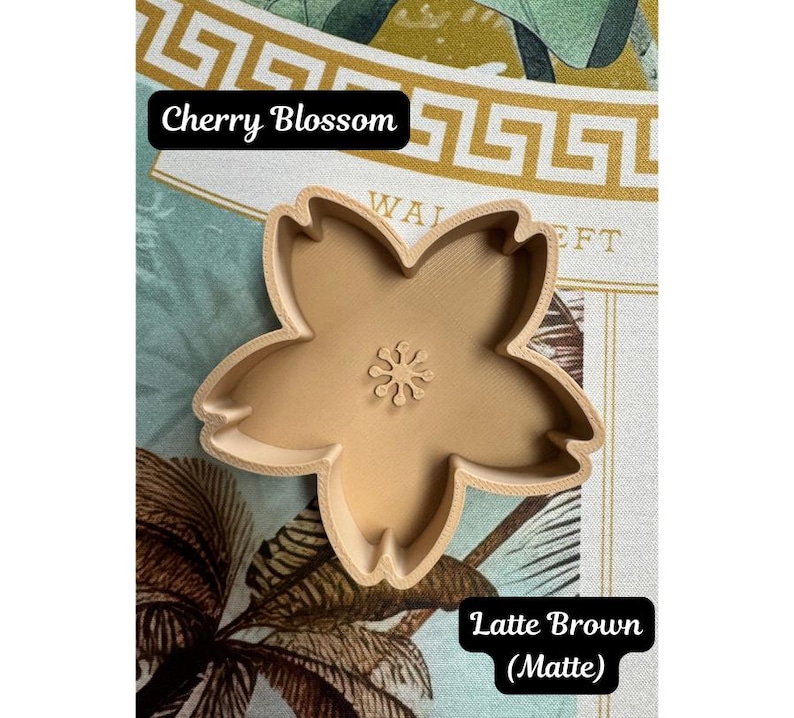 May include: A matte latte brown cookie cutter shaped like a five-petaled cherry blossom flower. The cutter features a central stamens detail and has a 3D printed texture. The text Cherry Blossom and Latte Brown (Matte) is visible in the image.