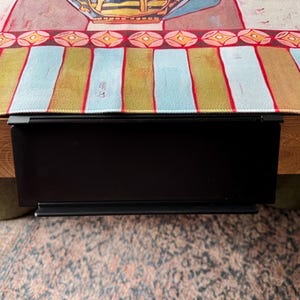 Puede incluir: Un estante de metal negro mate con abrazadera fijado al borde de una mesa de madera. El estante presenta una superficie rectangular plana con un peque&ntilde;o borde elevado en la parte inferior, colocado debajo de un camino de mesa de tela a rayas.