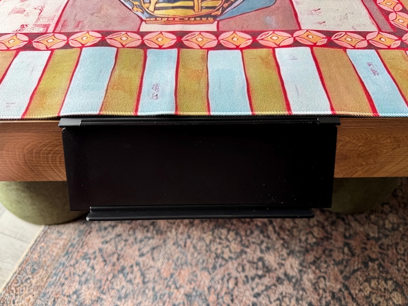 Puede incluir: Un estante de metal negro mate con abrazadera fijado al borde de una mesa de madera. El estante presenta una superficie rectangular plana con un peque&ntilde;o borde elevado en la parte inferior, colocado debajo de un camino de mesa de tela a rayas.