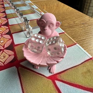 May include: A light pink 3D-printed monkey figurine holding a small, shallow, circular dish. Two transparent, six-sided dice with white pips are resting inside the dish. The monkey has a smooth, matte finish and is positioned upright.