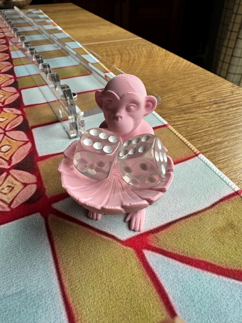 May include: A light pink 3D-printed monkey figurine holding a small, shallow, circular dish. Two transparent, six-sided dice with white pips are resting inside the dish. The monkey has a smooth, matte finish and is positioned upright.