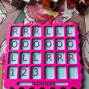 May include: A bright pink plastic Mahjong score card with a scalloped edge. The grid features four rows of rectangular slots. The top three rows contain the letters R, L, O, and the bottom row displays the numbers 1, 2, and 3. The word MAHJONG is embossed at the bottom center.