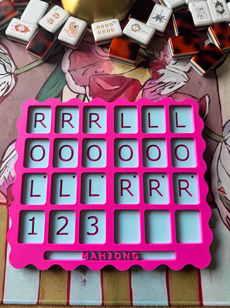 May include: A bright pink plastic Mahjong score card with a scalloped edge. The grid features four rows of rectangular slots. The top three rows contain the letters R, L, O, and the bottom row displays the numbers 1, 2, and 3. The word MAHJONG is embossed at the bottom center.