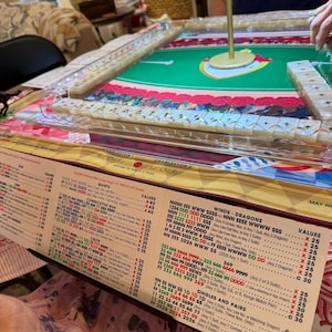 Puede incluir: Un juego de Mah Jong con una superficie de juego de fieltro verde y un poste central dorado, rodeado de filas de fichas blancas con s&iacute;mbolos de estrellas. Una tarjeta de puntuaci&oacute;n impresa con columnas de n&uacute;meros y texto detalla las reglas.