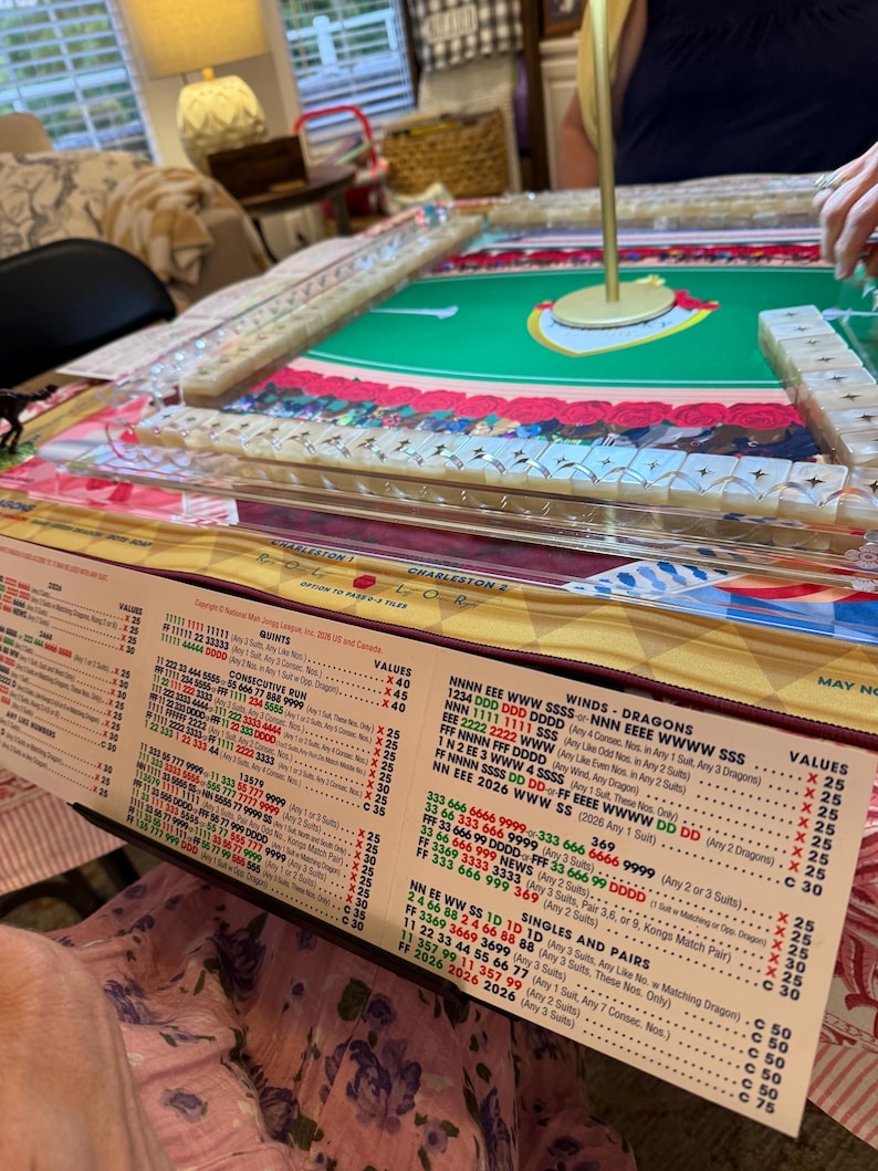 Puede incluir: Un juego de Mah Jong con una superficie de juego de fieltro verde y un poste central dorado, rodeado de filas de fichas blancas con s&iacute;mbolos de estrellas. Una tarjeta de puntuaci&oacute;n impresa con columnas de n&uacute;meros y texto detalla las reglas.