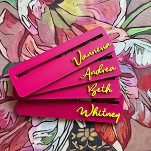 May include: Four rectangular hot pink bookmarks stacked on top of each other. Each bookmark features a horizontal slot and a name in yellow cursive script: Vannessa, Andrea, Beth, and Whitney. The bookmarks are made of a solid material.