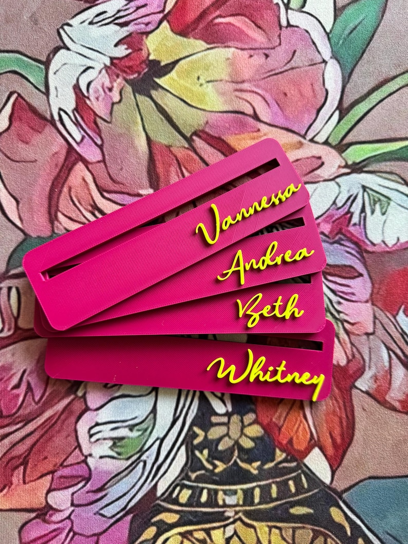 May include: Four rectangular hot pink bookmarks stacked on top of each other. Each bookmark features a horizontal slot and a name in yellow cursive script: Vannessa, Andrea, Beth, and Whitney. The bookmarks are made of a solid material.