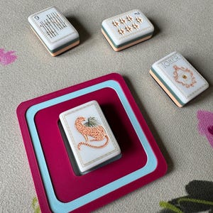 May include: A set of playing cards with a cheetah illustration, a card with the word "JOKER", and other cards with numbers and symbols. The cards are white with a peach and teal border, and are on a magenta and teal tray.