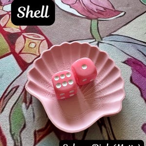 May include: A matte sakura pink shell-shaped dish containing two pink dice with white pips. The dish features a textured, ridged surface mimicking a seashell. Text on the image reads Shell and Sakura Pink (Matte) in black and white boxes.