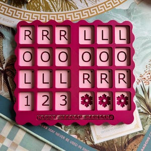 May include: A bright pink, scalloped-edge Mahjong tile rack featuring a grid of sixteen square cutouts. The rack displays the letters R, L, O, and the numbers 1, 2, 3, along with flower icons. The text MOUNT VERNON MAHJONG is embossed at the bottom.